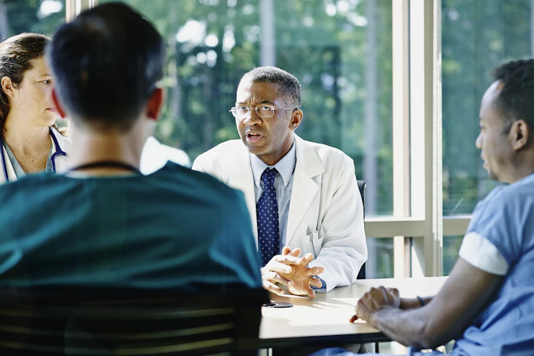 A doctor in a white coat and tie sits at a table talking to other doctors and nurses, with trees seen through the window.