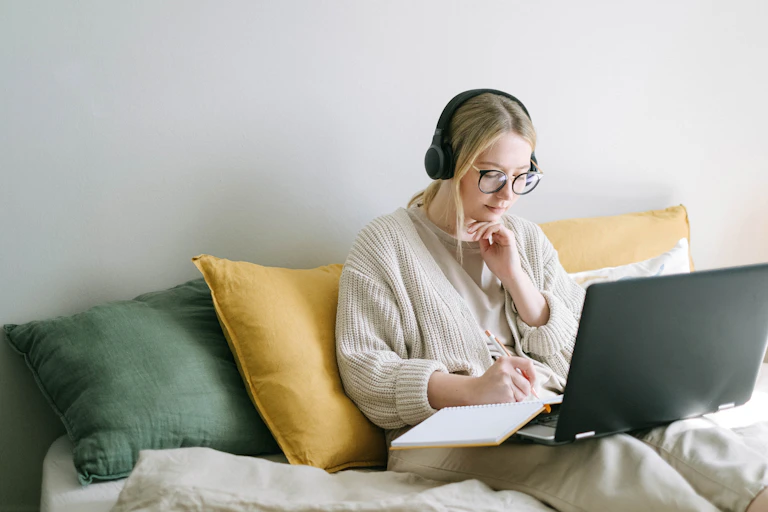 A young woman sits on a bed, working on a laptop.