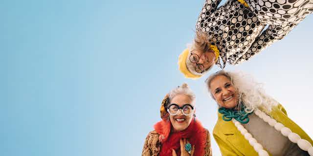 Tres señoras mayores con ropas de colores miran a una cámara situada en el suelo mientras sonríen.