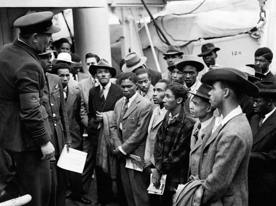 Windrush passengers welcomed by officials from the Colonial Office wearing smart hats and double breasted suits. 
