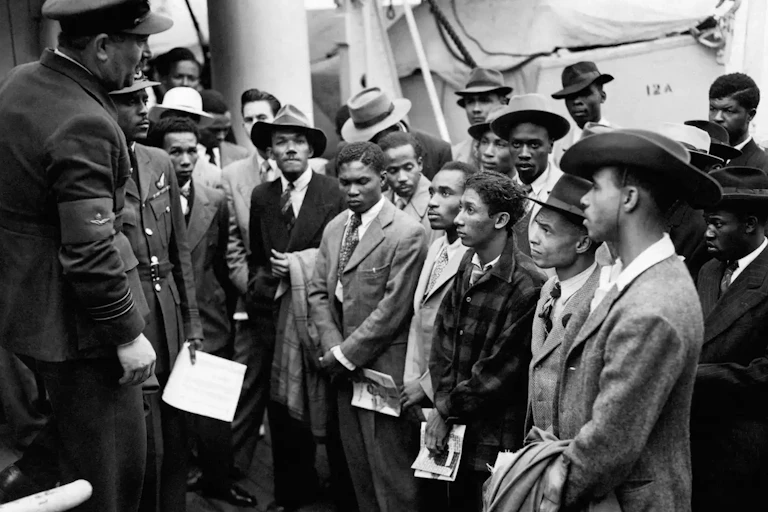 Windrush passengers welcomed by officials from the Colonial Office wearing smart hats and double breasted suits.