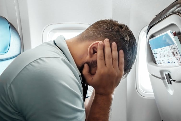 A man sat in front of an airplane window with his head in his hands.