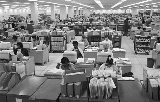 A black and white photo of people working in a vast office using blocky 1980s-style computers.