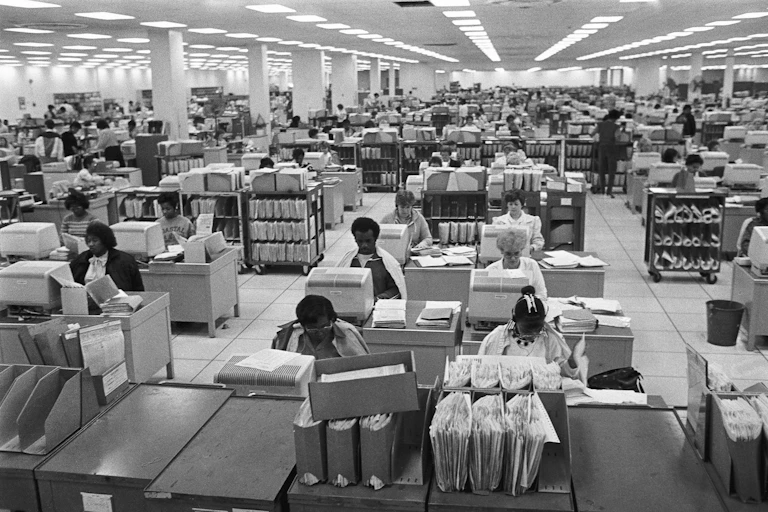 A black and white photo of people working in a vast office using blocky 1980s-style computers.