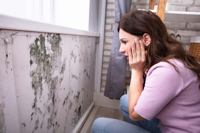 A young woman squats down and looks in horror at a wall covered in black mould