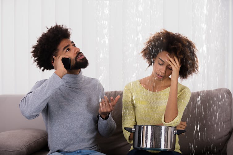 A young couple sits on a couch under a leaking ceiling, the man is on the phone and the woman is holding a saucepan in her lap to collect water