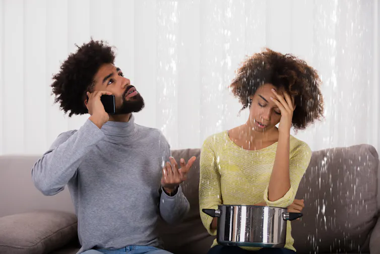 A young couple sits on a couch under a leaking ceiling, the man is on the phone and the woman is holding a saucepan in her lap to collect water