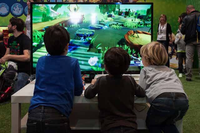 Tres niños pequeños juegan a un videojuego con consola en una habitación llena de gente.