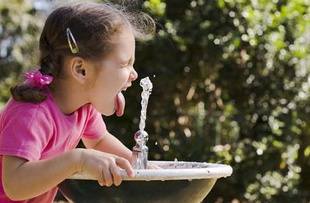 A young girl sticking her tongue out in a drinking fountain on a hot day