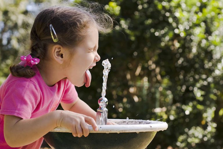 A young girl sticking her tongue out in a drinking fountain on a hot day