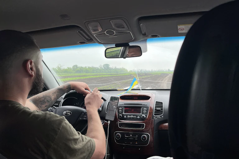 Photo prise de la banquette arrière d'une voiture en marche. Un homme est au volant, un drapeau ukrainien sur le tableau de bord. La voiture roule dans la campagne.