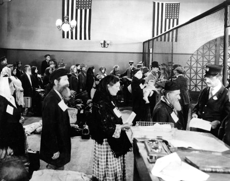 A black and white photo of long lines of people with luggage in an old-fashioned arrival hall.