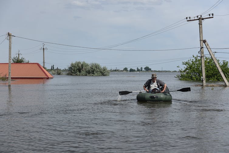 A man is seen using oars to power his inflatable small boat along a flooded street.