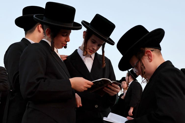 Four teenage boys in black coats and black, broad-brimmed hats study a book while standing outside.
