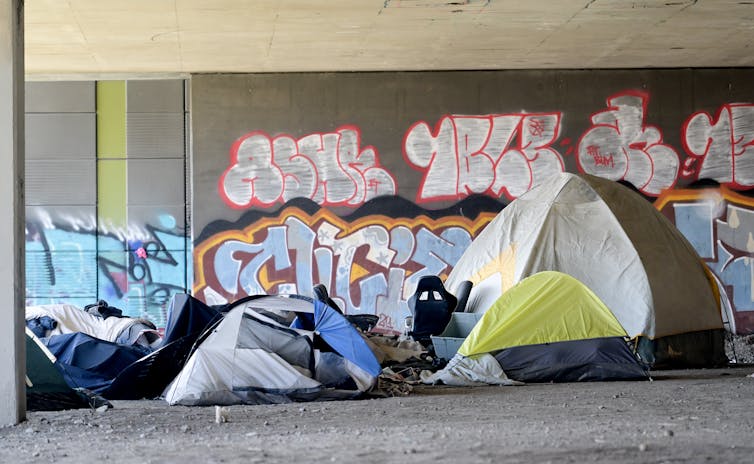 Tents beneath an overpass. Graffiti is seen on the wall behind them.