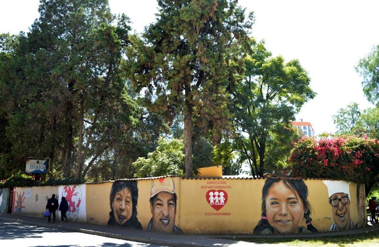 Murals of children's faces on a yellow wall below tall trees.