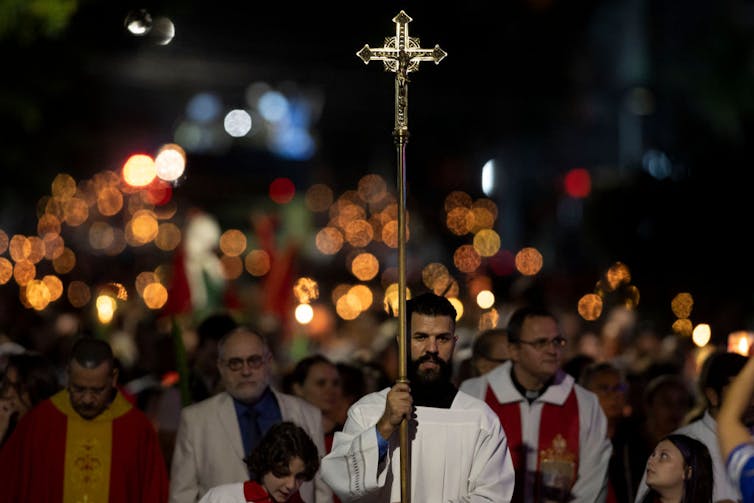 A religious procession walking behind a man holding a tall metal cross at nighttime.