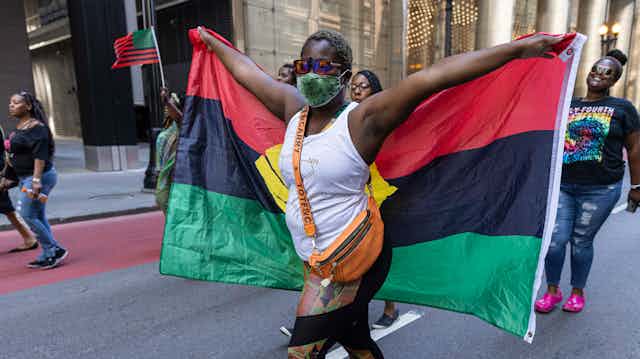 A woman seen holding a red, black and green striped flag with her arms outstretched at a rally.