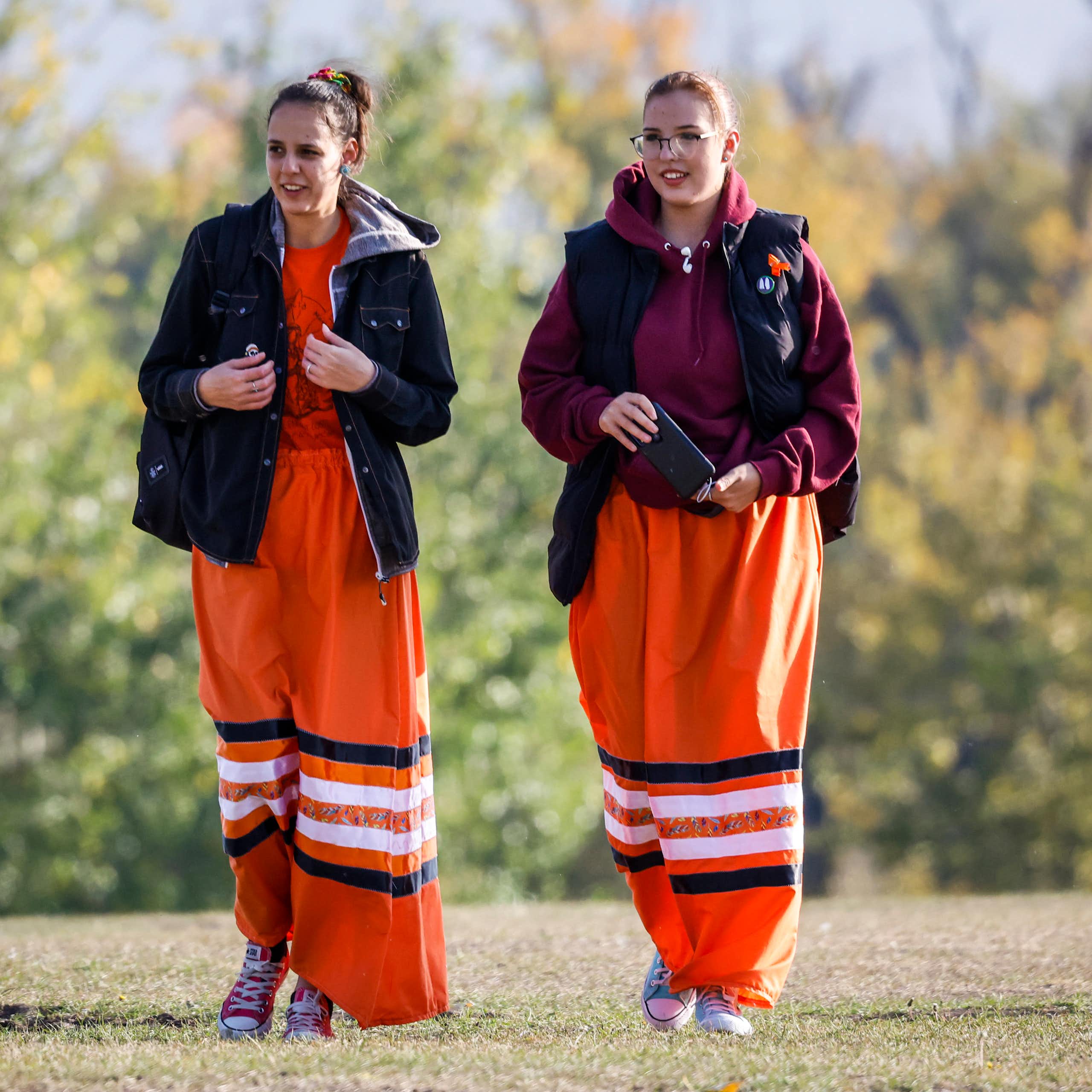 Two young women walk side by side wearing orange ribbon skirts and black jackets in an open green area