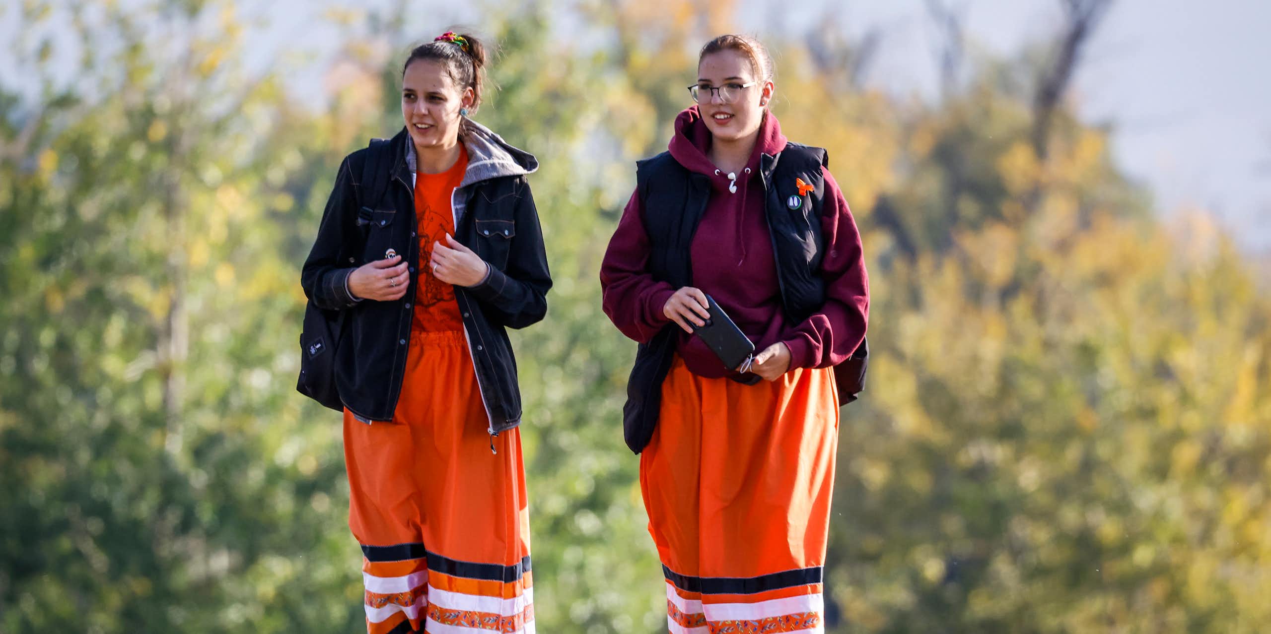 Two young women walk side by side wearing orange ribbon skirts and black jackets in an open green area