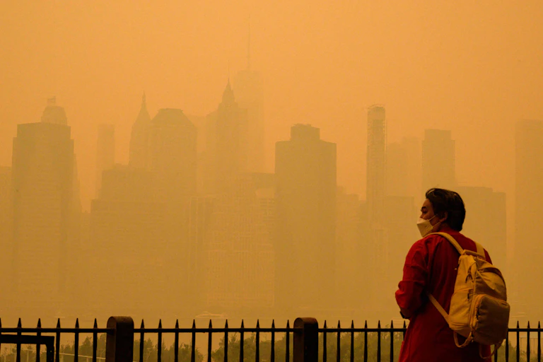 A person wearing a facemask stands at a railing looking out across the water at the NYC skyline in the orange haze of wildfire smoke. One World Trade Center is barely visible.