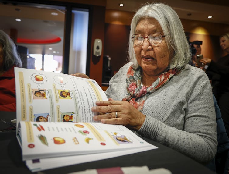 Elder woman wearing grey shirt and colorful scarf reading a children's book.