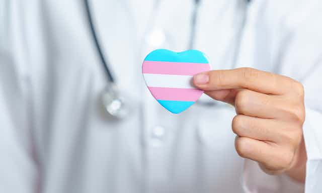 A person in a white coat and stethoscope out of focus holding a blue, pink and white striped heart in the foreground