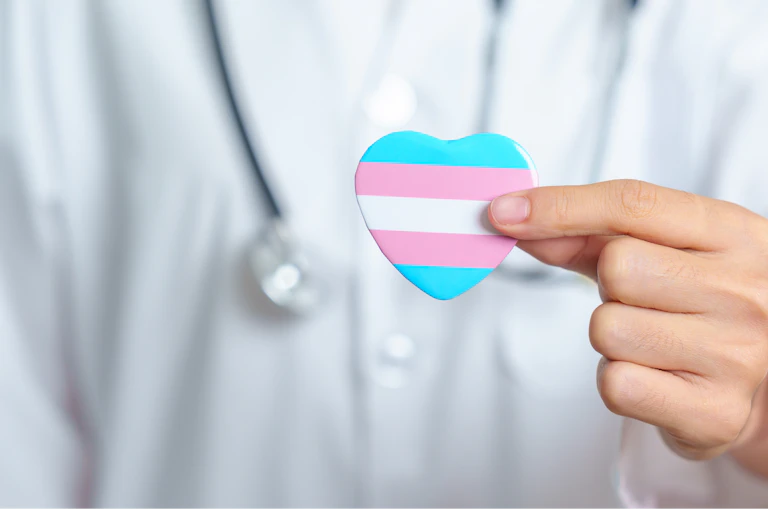 A person in a white coat and stethoscope out of focus holding a blue, pink and white striped heart in the foreground