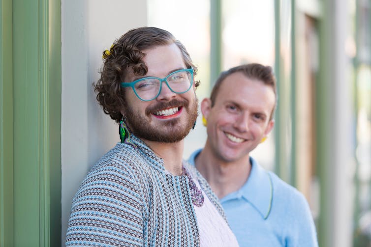 A person with a beard and glasses wearing a striped shirt smiling and a second person out of focus