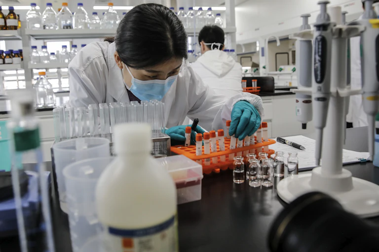 A Chinese woman in COVID mask leans over a bench inspecting the contents of a test tube.
