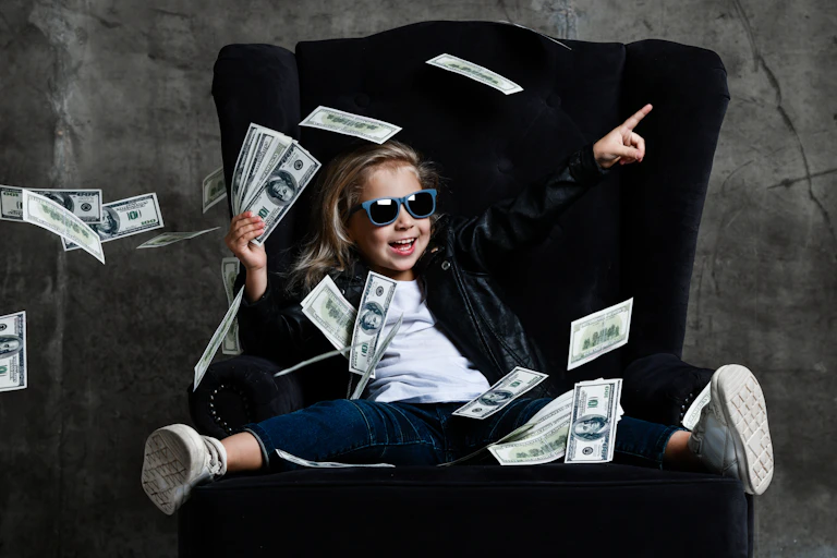 Una niña sentada en un sillón tiene un montón de billetes en la mano mientras más billetes vuelan a su alrededor.