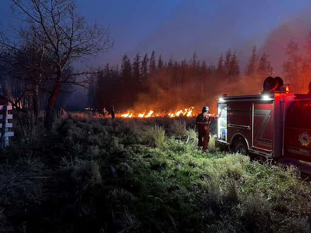A parked fire engine at dusk with a firefighter stood next to it and fire in the distance.