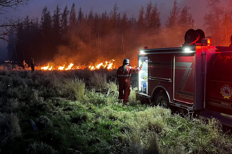 A parked fire engine at dusk with a firefighter stood next to it and fire in the distance.