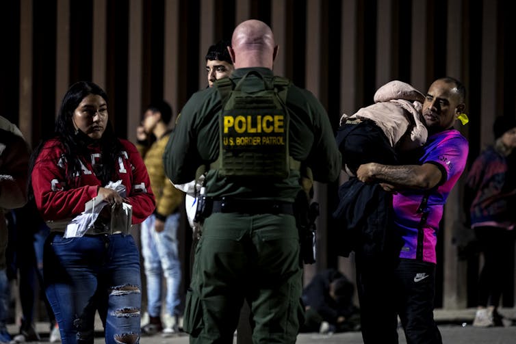 Um policial careca de costas fica diante de pessoas carregando crianças adormecidas.
