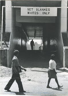 Two people of colour pass an entrance to a public facility that has a large sign above it reading 