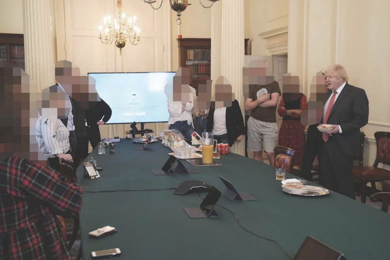Boris Johnson standing with people whose faces have been pixellated. They are in a government room around a table with refreshments on it.