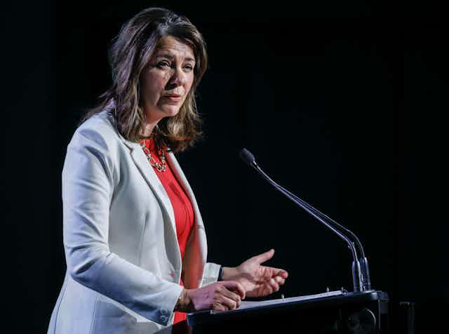 A dark-haired woman in a grey jacket and red shirt speaks at a podium with a microphone in front of her.