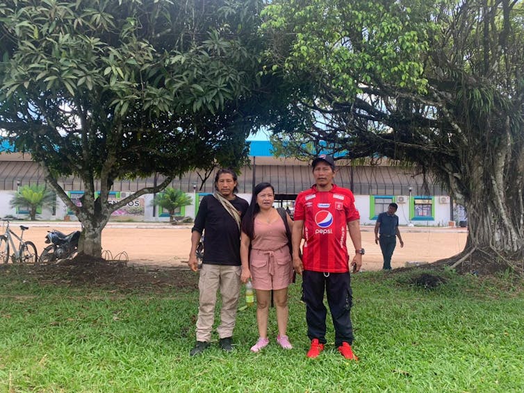 Three Indigenous people in western clothes stood under trees in front of a wide building.