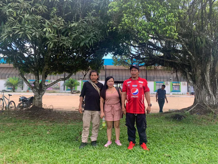 Three Indigenous people in western clothes stood under trees in front of a wide building.