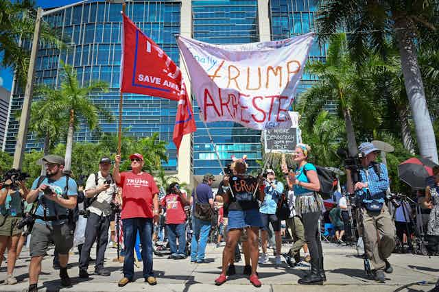 People stand together outside of a large, glass building, holding signs that say 'Trump arrested' and also 'Trump, save America!'