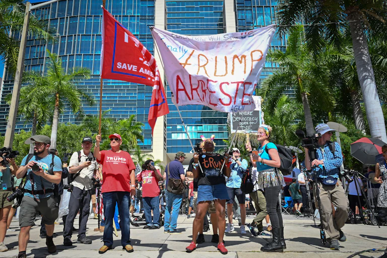 People stand together outside of a large, glass building, holding signs that say 'Trump arrested' and also 'Trump, save America!'