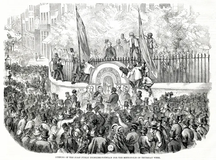 Archive illustration showing a drinking fountain surrounded by a crowd of people, mostly men in top hats, with children climbing the fence around the fountain