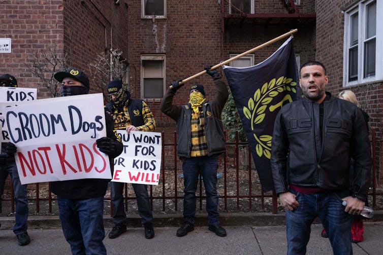 Varios hombres se paran afuera de un edificio de ladrillo rojo y sostienen carteles que dicen cosas como 'Prepara perros, no niños'.
