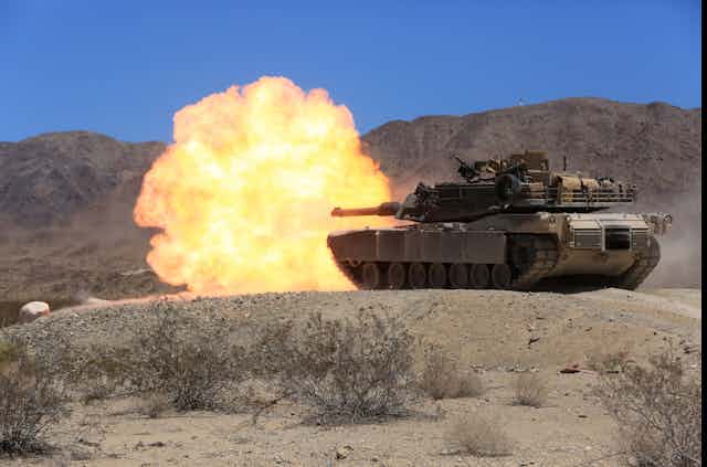 A tank on a sandy range with a large explosion coming from its main gun
