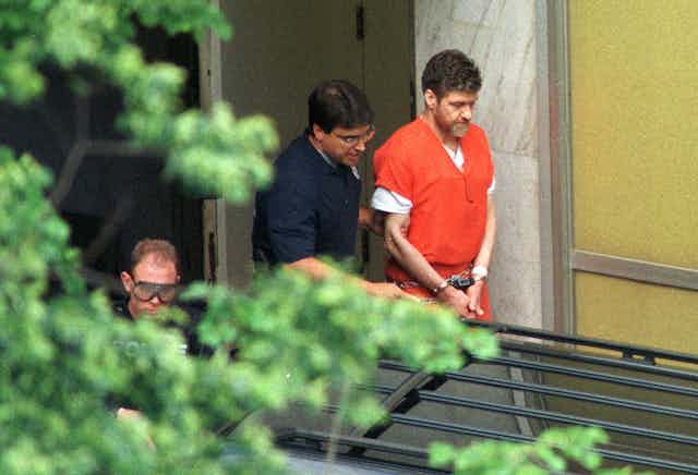 Handcuffed man with beard in an orange jumpsuit being led by another man.