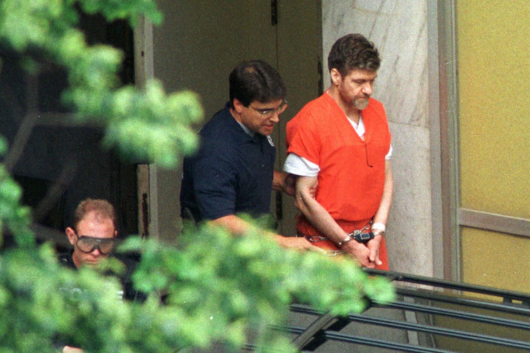 Handcuffed man with beard in an orange jumpsuit being led by another man.