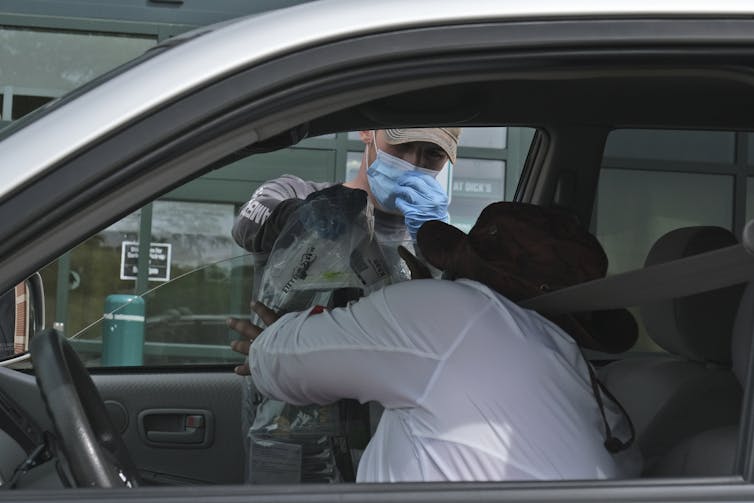 A person wearing a medical face mask passes someone a grocery bag through their open car window