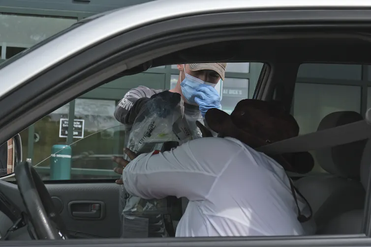 A person wearing a medical face mask passes someone a grocery bag through their open car window