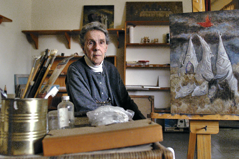An elderly Carrington sat in her studio, with a painting behind her and brushes to her side.