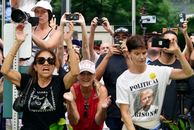 Three people protesting as part of a crowd. One wears a t-shirt with a photo of Donald Trump that says 'political prisoner.'